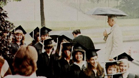 Graduates walk in a line with a man holding an umbrella behind them.