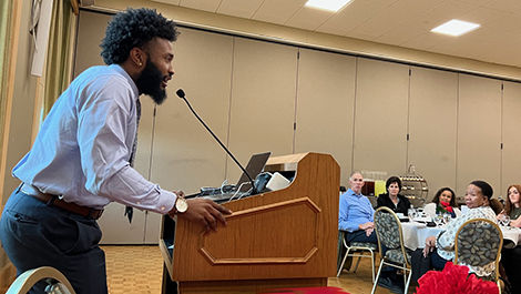 Standing at a podium, Joseph Clark speaks to people seated at tables.