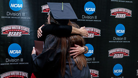 A graduate hugs a coach in front of a BSU athletics backdrop.