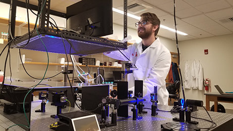 Josh Watts, wearing a white lab coat, looks at a monitor above optical tweezers.