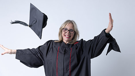 Lesly Freed, wearing her black graduation gown, tosses her cap in the air in front of a white background.