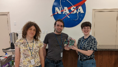 Three people stand holding a circuit board in front of a NASA sign