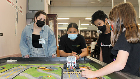 A BSU student helps fifth graders test a robot on a mat.