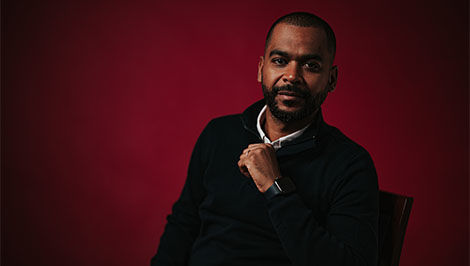 portrait photo of Harold Tavares sitting in a chair in front of a red backdrop