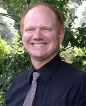 Jon Amon smiling with receding short red hair wearing navy blue button down shirt with gray blue tie and trees in background