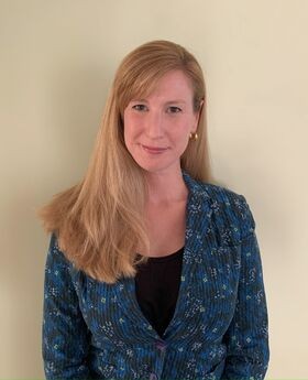 Maria Armour smiling with long dirty blonde hair parted on the side and wearing a black top under a blue, black, white and yellow print blazer