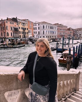 Ashley Hartsfield smiling with long dirty blonde hair and wearing a long sleeve black top while standing on a stairway overlooking a canal in Venice, Italy