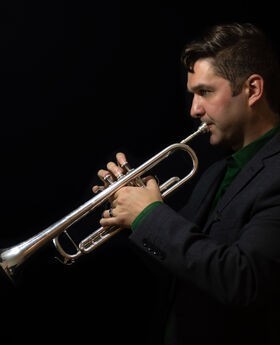 Dr. Spencer Aston with short dark brown hair playing trumpet while looking at sheet music. He is wearing black on a black background