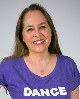 Ann Biddle smiling with long brown hair and wearing a purple t-shirt that says "DANCE" on it