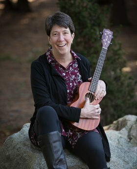 Paula Bishop sitting on a rock smiling with short dark brown hair, holding a ukulele and wearing black leggings and tall fashion boots and a black open sweater over a long maroon button up blouse