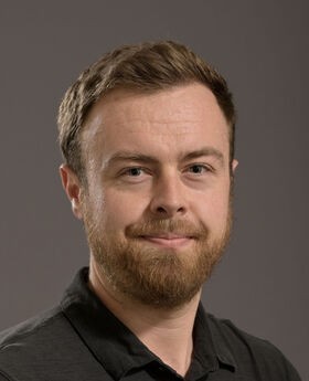 Dr. Myles Bittner smiling with short reddish brown hair, mustache and beard. He is wearing a black polo shirt