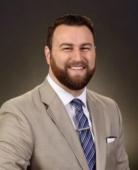 Dr. Lance Brooks smiling with short brown hair, beard and mustache. He is wearing a tan suit jacket over a white button down shirt and blue tie