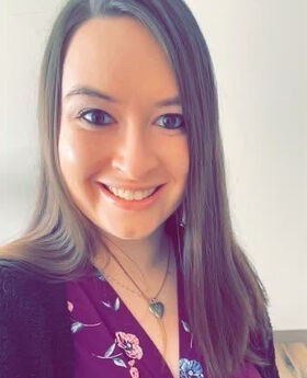 Christine Camara smiling with long straight brown hair and wearing a purple wrap dress with a black sweater