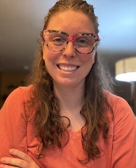 Tori Cameron smiling with long wavy light brown hair and wearing print rim glasses and an orange v-neck top