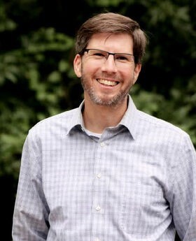 Ben Carson with short brown hair graying at the temples, brown and gray mustache and beard wearing glasses and a light blue button down shirt