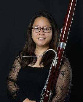 Debbie Chen smiling with long dark brown hair and wearing black rim glasses and a black dress with sheer sleeves. She is holding a bassoon.