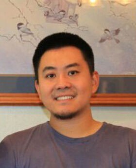 Dr. Xiangfei Chen smiling with short black hair and a short goatee. He is wearing a dark gray t-shirt/jersey with framed artwork behind him