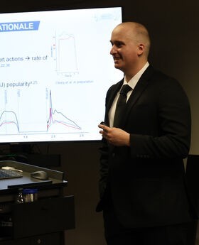 Dr. CJ Cleary teaching next to a projector screen and smiling while wearing a black suit and tie with a tan button down shirt