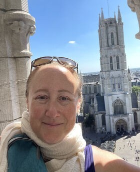 Dr. Jodi Cohen smiling with her brownish/red hair pulled back and sunglasses on top of her head. She is wearing a beige linen scarf and sleeveless purple top, standing on a balcony with a gothic church in the background