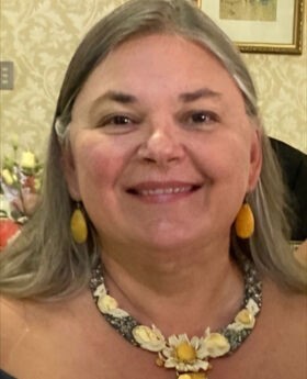Veronica Cote smiling with medium length gray hair and wearing a black off the shoulder dress with gold colored earrings and necklace