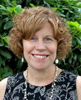 Diane Crowley smiling with medium length curly light brown hair and wearing a sleeveless black with white print top