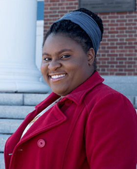 Daniela Belice standing on the steps of Boyden Hall smiling at the camera with her black hair pulled up in a bun and wearing a red coat with lapel and buttons