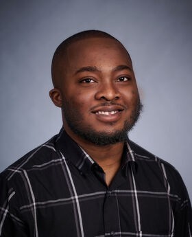 Dr. Edwin Dartey smiling with shaved black hair and a short mustache and beard. He is wearing a black button down shirt with large gray box stripes