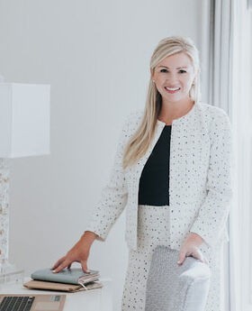 Dr. Kaitlin Lambert Donahue smiling with long straight blondish hair. She is standing next to a desk in a white room wearing a white suit with small black polka dots and a black blouse