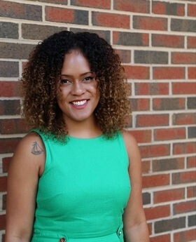Karina Dos Santos standing in front of a brick wall smiling with medium length curly brownish black hair and wearing a sleeveless green top.