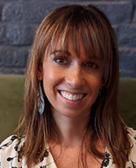 headshot photo of Cynthia Drake smiling with medium length light brown hair and wearing a white v-neck blouse with black flowers print