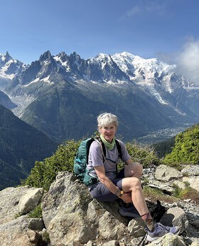Dr. Susan Eliason sitting on top of a mountain smiling with short gray hair wearing blue shorts, t-shirt, backpack and hiking sneakers