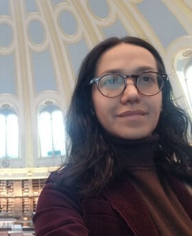 a selfie photo of Dr. Carolina Freitas inside an architectural dome decorated in a light blue, cream and gold design. Dr. Freitas has long dark brown wavy hair and is wearing brown rim glasses and a cranberry colored corduroy blazer over a brown turtleneck top.