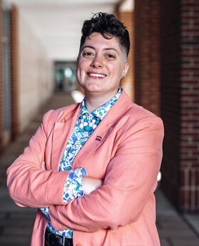 Dr. Reihonna Frost-Calhoun smiling with short dark brown curly hair wearing a salmon colored blazer with rainbow flag pin on it and blue and white print button down shirt under it