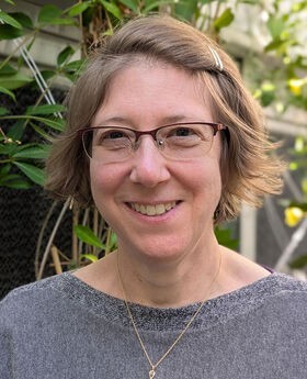 Dr. Laura K. Gross in the BSU Greenhouse smiling with short light brown hair and wearing brown rim glasses and a gray boat neck top.