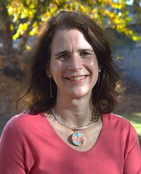 Dr. Lynn Hansberry Mayo smiling with medium length dark brown wavy hair and wearing a salmon v-neck top