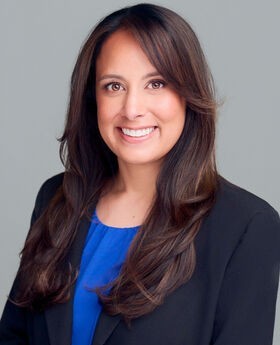 Christina Hodel smiling with long wavy dark brown hair and wearing a navy blazer over a light blue blouse