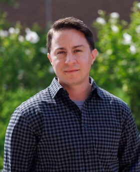 Dr. Asa Iacobucci standing outside in front of greenery with short brown hair wearing a black and purple check button up shirt