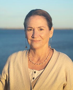 headshot photo of Dr. Margaret Kjelgard with her brown hair pulled back, smiling and wearing a beige v-neck sweater with ocean in the background