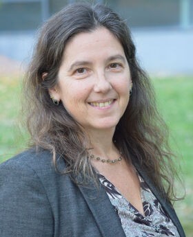 Brenda Kozuch smiling with dark brown wavy long hair wearing a charcoal gray blazer over a v-neck gray and black top