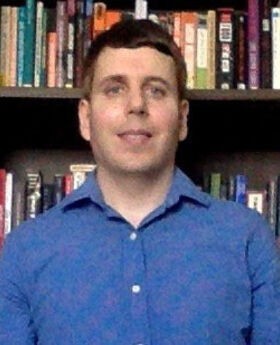Brian Lepine with short brown hair and blue eyes wearing a blue button down shirt and standing in front of a bookcase