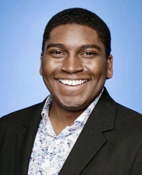 Dr. Niles Mattier smiling with short black hair and wearing a black blazer over a white and blue print button down shirt