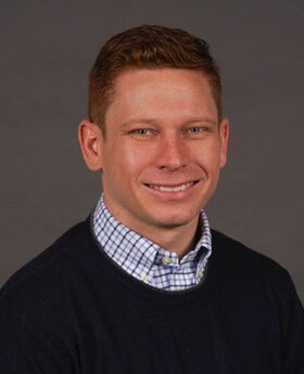 Dr. Ted McClain smiling with short wavy reddish brown hair and wearing a navy blue sweater over a white shirt with blue stripes on it