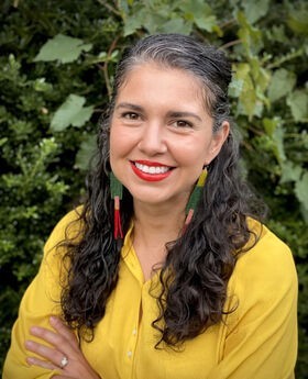 Dr. Alicia Mendez smiling in front of greenery with long graying dark brown hair pulled back on top and wearing a dark yellow button up blouse and long colorful earrings
