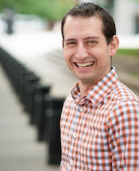 Dr. Seth Meyer looks at the camera and smiles in a red and blue checkered shirt outside