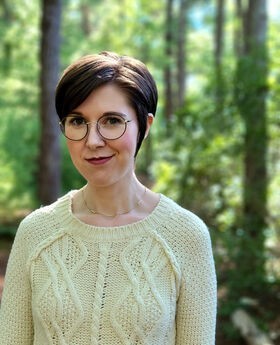 Dr. Sarah Fawn Montgomery standing in front of a wooded background with short dark brown hair and wearing round black rim glasses and a pale yellow cable knit sweater