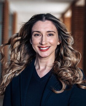 Patricia Morchel smiling with long wavy brown hair wearing a navy blue blazer with navy blue v-neck top under it