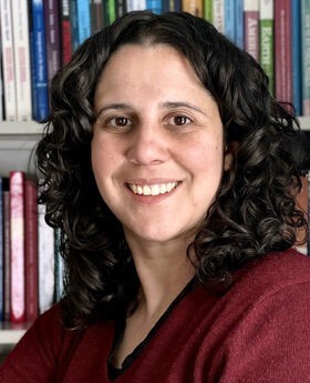 Dr. Rachel Navarre smiling in front of a bookcase with long curly dark brown hair and wearing a v-neck maroon sweater