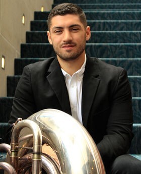 Jordan Oliveira sitting on a carpeted stairway with his tuba. He has short dark brown hair, mustache and beard and is wearing a black suit over a white button down shirt