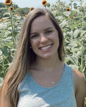 Olivia Dean, '22, smiling with long straight light brown hair and wearing a light blue tank top with sunflowers in the background