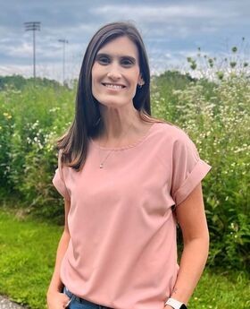 Dr. Kerri Olore standing in front of outdoor flowers and greenery smiling with long straight dark brown hair and wearing a short sleeve pink blouse and jeans